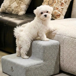 May include: A small, fluffy white dog stands on a two-step gray pet staircase. The staircase has a textured fabric and a logo on the side. The dog is looking at the camera. The background includes a black sofa and decorative pillows.