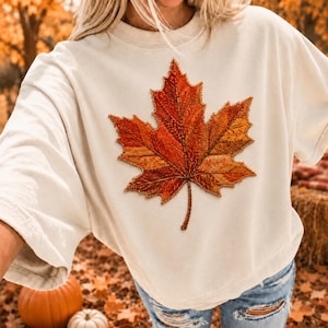 Peut inclure: Un t-shirt à col rond blanc cassé arborant un grand motif de feuille d'érable brodé et détaillé dans des tons orange, rouge et doré. Le motif de la feuille est centré sur la poitrine et présente des points de couture complexes sur tout le feuillage.