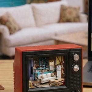 May include: A small red television with a black screen showing a detailed interior scene. The TV has knobs and buttons and is placed on a wooden surface. The scene inside the TV appears to be a living room, with a sofa.