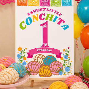 May include: A white rectangular sign featuring the text A SWEET LITTLE CONCHITA 1 TURNS ONE in colourful letters. The design includes illustrations of five concha sweet breads in pink, blue, yellow, and brown, accented by floral motifs and banners.