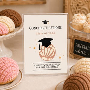 May include: A clear acrylic sign with the text "CONCHA-TULATIONS Class of 2026" and a graduation cap illustration. The sign is surrounded by concha pastries and cupcakes, with a small chalkboard sign that reads "PAN DULCE bar".