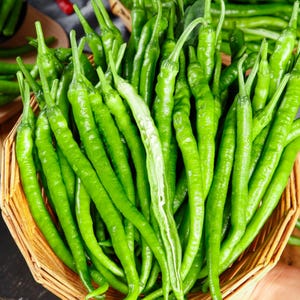 Può includere: Un cesto intrecciato pieno di numerosi peperoncini verdi lunghi e sottili. I peperoncini hanno una buccia liscia e lucida con gambi sottili. Un peperoncino al centro è tagliato a metà per il lungo, rivelando l'interno chiaro e i semi.