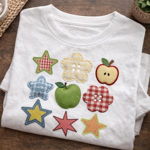 May include: White t-shirt decorated with colorful fabric appliqués of stars, flowers, and apples. The appliqués feature various patterns and textures, including gingham and floral prints. A charming, handmade design.