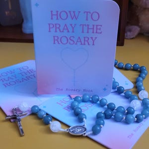 May include: A folded card with a gradient background featuring the text HOW TO PRAY THE ROSARY and a heart-shaped rosary illustration. A blue beaded rosary with a silver crucifix and a central medal rests on the card, which also displays The Rosary Nook.