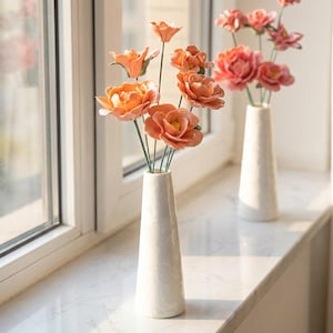 May include: Two white ceramic vases with orange and pink flowers. The flowers have green stems and are arranged in a decorative display. The vases are on a white windowsill near a window.