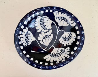 Op de afbeelding: Een ronde keramische schaal met een diep donkerblauw glazuur en een centraal handgeschilderd ontwerp van vijf witte bloemen met gestructureerde bloemblaadjes. De rand is versierd met een herhalend patroon van witte vierkantjes en blauwe stippen.