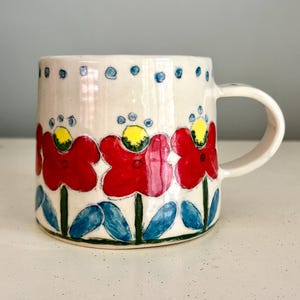 May include: A white ceramic mug featuring a hand-painted floral pattern. The design includes red flowers with yellow centers, green stems, and blue leaves. A row of small blue dots encircles the top rim of the mug, which has a side handle.