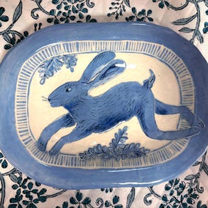 Puede incluir: Un plato ovalado de cerámica con un conejo azul pintado a mano en pose de salto sobre un fondo color crema. El plato tiene un borde ancho azul con un patrón de líneas verticales y detalles decorativos de follaje azul en la parte superior e inferior.