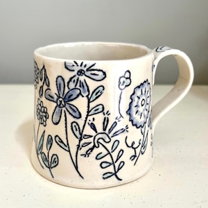 May include: A ceramic mug with a white base and a blue floral design. The flowers and leaves are hand-drawn with black outlines. The mug has a curved handle and is suitable for hot or cold beverages.