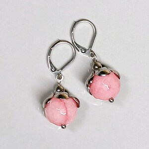 May include: A pair of dangle earrings featuring light pink, spherical beads. The beads are encased in a silver-colored metal setting and attached to lever-back closures. The earrings are displayed on a white background.
