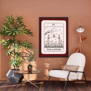 以下が含まれることがあります： 「THE LOVERS」のテキストが入った、2体の骸骨が花を持っている白黒イラストの、額入りタロットカードプリント。プリントには赤い縁取りがあり、桃色の壁に飾られています。