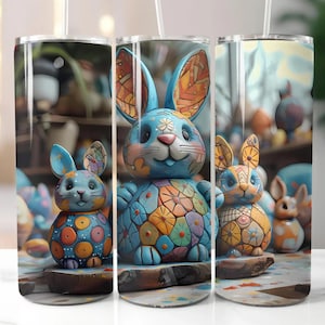 Op de afbeelding: Drie roestvrijstalen bekers met een kleurrijk paashaasontwerp. De konijnen zijn blauw met oranje, gele en groene accenten. De bekers hebben een doorzichtig rietje en staan tegen een wazige achtergrond van paasdecoraties.