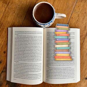 May include: An open book with colorful book-shaped bookmarks, a cup of coffee, and a blue and white patterned mug on a wooden surface. The bookmarks are stacked, creating a vibrant display.