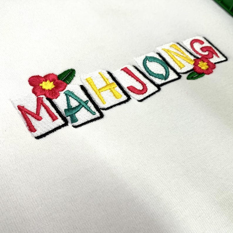 Puede incluir: Sudadera blanca con la palabra "MAHJONG" bordada en rojo, verde, amarillo y negro. Las letras est&aacute;n sobre rect&aacute;ngulos blancos individuales con bordes negros. Flores rojas con hojas verdes bordadas a ambos lados.