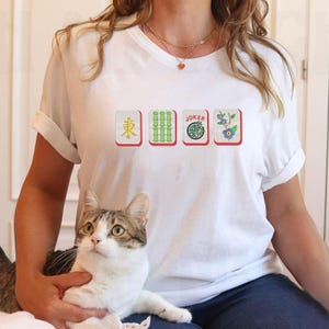 May include: White t-shirt featuring four Mahjong tile designs. The tiles include a character, bamboo, a "JOKER" tile, and a floral design. The shirt is worn by a person holding a cat.