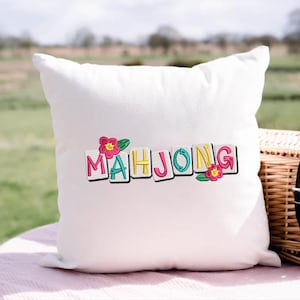 Puede incluir: Coj&iacute;n cuadrado blanco con la palabra "MAHJONG" bordada en letras coloridas. Las letras est&aacute;n delineadas en negro y adornadas con flores rosas y hojas verdes. El coj&iacute;n est&aacute; sobre una superficie rosa.