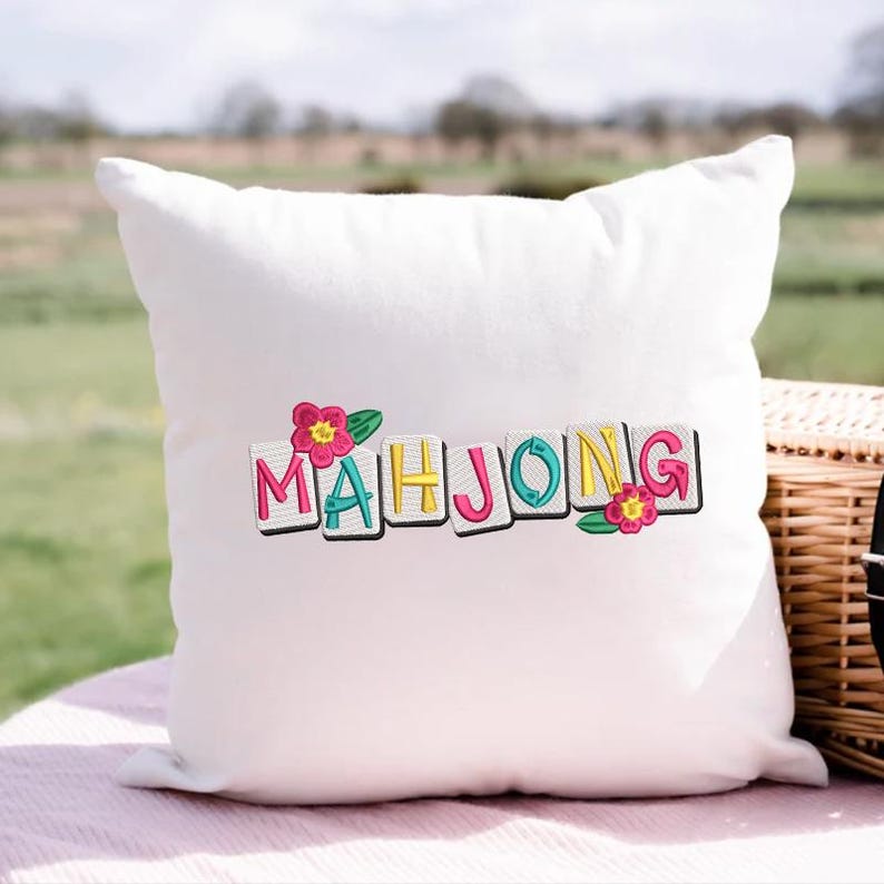Puede incluir: Coj&iacute;n cuadrado blanco con la palabra "MAHJONG" bordada en letras coloridas. Las letras est&aacute;n delineadas en negro y adornadas con flores rosas y hojas verdes. El coj&iacute;n est&aacute; sobre una superficie rosa.