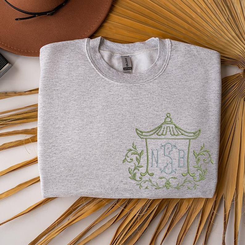 May include: A light gray crewneck sweatshirt featuring an embroidered design on the lower right side. The embroidery depicts a light green pagoda-style structure framing the letters NSB in a blue script font, surrounded by light green leafy vines.