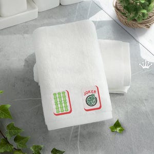 May include: White bath towel with two embroidered Mahjong tiles and the word "JOKER". The tiles are green and red. The towel is folded and stacked with another white towel. The background is a light gray surface.