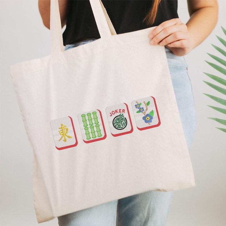 May include: Off-white tote bag featuring four Mahjong tile designs. The tiles include the East wind, bamboo, a Joker tile, and a floral tile. The bag has a simple design with a focus on the colorful tile graphics.