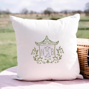 May include: A square white throw pillow featuring an embroidered design in the center. The embroidery depicts a green pagoda-style roof structure framing a light blue monogram with the letters N, S, and B, surrounded by green leafy scrollwork.