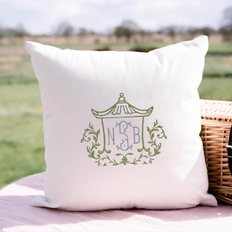 May include: A square white throw pillow featuring an embroidered design in the center. The embroidery depicts a green pagoda-style roof structure framing a light blue monogram with the letters N, S, and B, surrounded by green leafy scrollwork.