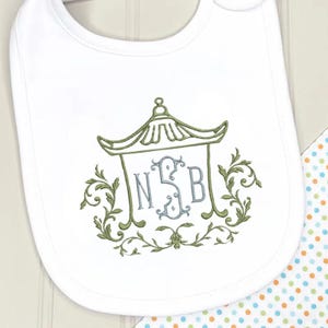 May include: A white baby bib featuring an embroidered green pagoda design surrounding a light blue monogram with the letters NSB. The embroidery is accented with green scrollwork and leaf motifs, centered on the fabric surface of the bib.