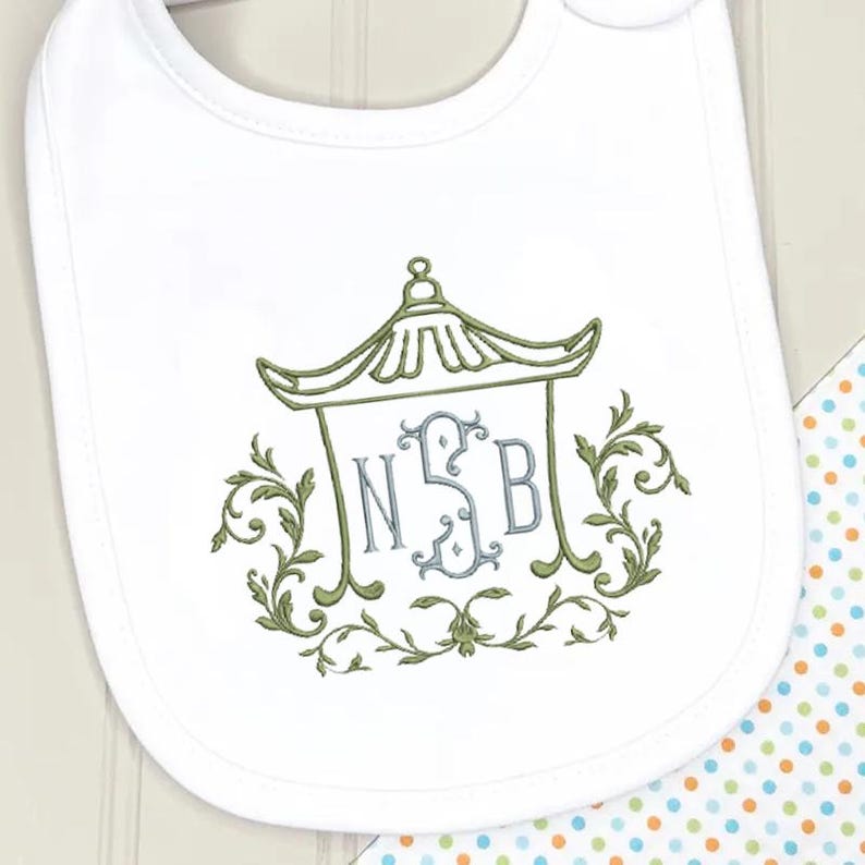 May include: A white baby bib featuring an embroidered green pagoda design surrounding a light blue monogram with the letters NSB. The embroidery is accented with green scrollwork and leaf motifs, centered on the fabric surface of the bib.