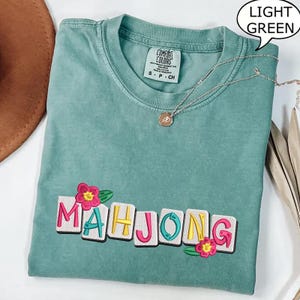 Puede incluir: Camiseta verde claro con la palabra "MAHJONG" bordada en letras coloridas y flores. La camiseta tiene cuello redondo y mangas cortas. Un collar plateado con un colgante est&aacute; drapeado sobre la camiseta.