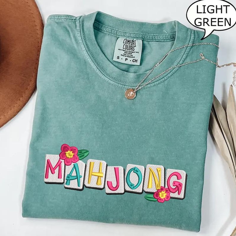 Puede incluir: Camiseta verde claro con la palabra "MAHJONG" bordada en letras coloridas y flores. La camiseta tiene cuello redondo y mangas cortas. Un collar plateado con un colgante est&aacute; drapeado sobre la camiseta.