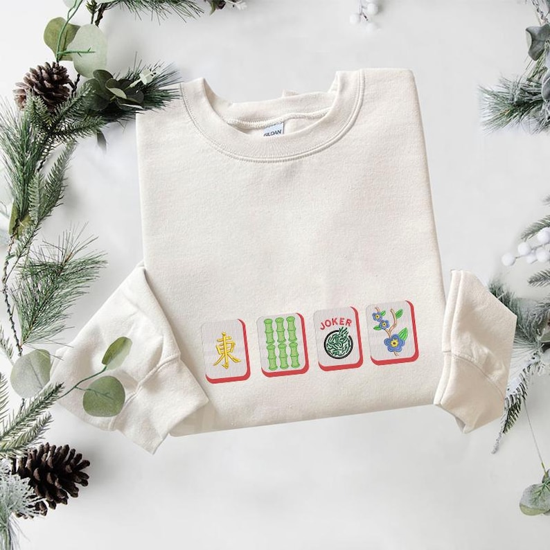 May include: Cream-colored sweatshirt featuring embroidered Mahjong tiles: East, Bamboo, Joker, and a floral tile. The design is in red, green, blue, and white. The sweatshirt has a crew neck and long sleeves.