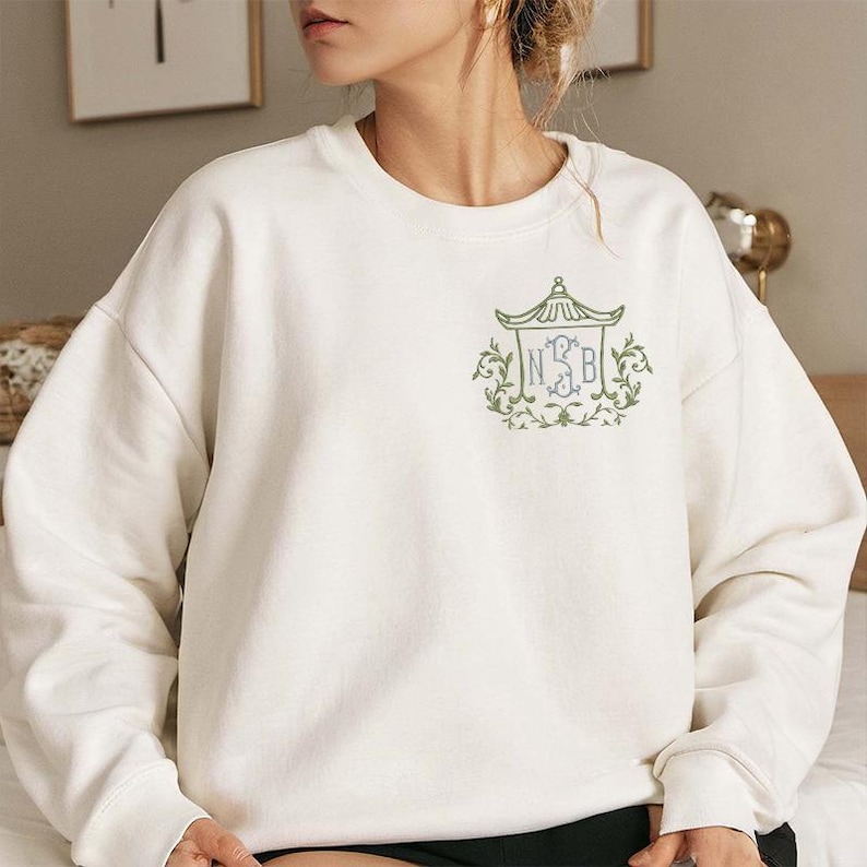 May include: A white long-sleeve sweatshirt featuring an embroidered design on the left chest. The embroidery depicts a green pagoda-style structure framing the letters NSB in blue, surrounded by green leafy vines and decorative scrollwork.
