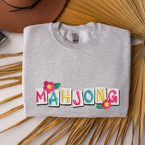 Puede incluir: Una sudadera gris claro con la palabra "MAHJONG" bordada en letras coloridas. Flores rosas y hojas verdes est&aacute;n bordadas a ambos lados de la palabra. La sudadera est&aacute; doblada sobre una superficie blanca.