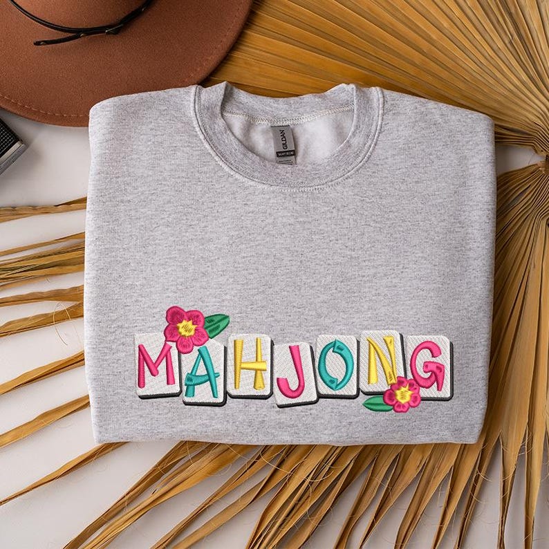Puede incluir: Una sudadera gris claro con la palabra "MAHJONG" bordada en letras coloridas. Flores rosas y hojas verdes est&aacute;n bordadas a ambos lados de la palabra. La sudadera est&aacute; doblada sobre una superficie blanca.