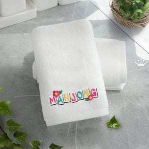 Puede incluir: Toalla de ba&ntilde;o blanca con la palabra "MAHJONG" bordada en letras coloridas y detalles florales. Otra toalla m&aacute;s peque&ntilde;a est&aacute; apilada al lado. El fondo es una superficie gris claro.