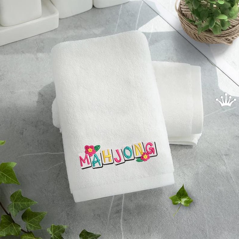 Puede incluir: Toalla de ba&ntilde;o blanca con la palabra "MAHJONG" bordada en letras coloridas y detalles florales. Otra toalla m&aacute;s peque&ntilde;a est&aacute; apilada al lado. El fondo es una superficie gris claro.