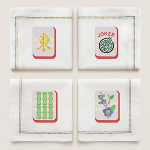 May include: Four white linen cocktail napkins with embroidered Mahjong tile designs. Each napkin features a different tile: "East" in gold, a "Joker" tile, green bamboo, and blue flowers. The napkins have a red border and a stitched edge.