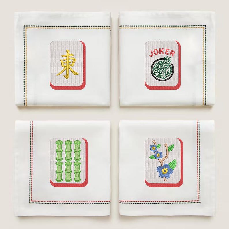 May include: Four white linen cocktail napkins with embroidered Mahjong tile designs. Each napkin features a different tile: "East" in gold, a "Joker" tile, green bamboo, and blue flowers. The napkins have a red border and a stitched edge.