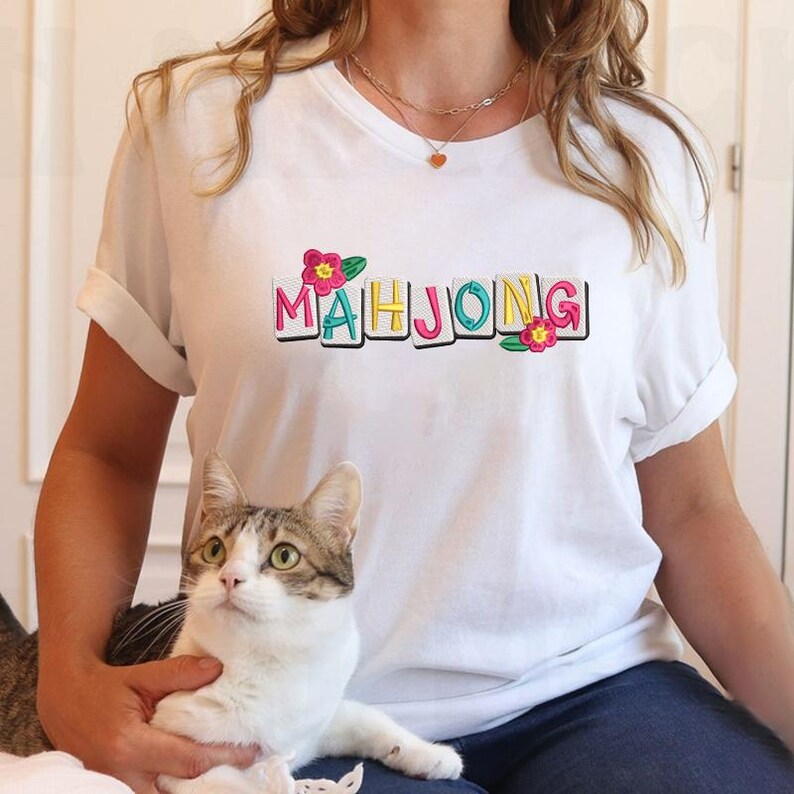 Puede incluir: Camiseta blanca con la palabra "MAHJONG" en letras de bloque coloridas, acentuada con flores rosas y amarillas. Un gato con ojos verdes y pelaje gris y blanco est&aacute; en primer plano. La camiseta tiene mangas enrolladas.