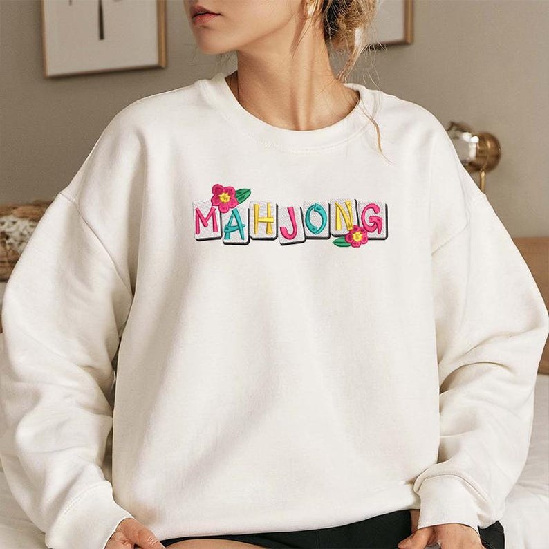 Puede incluir: Sudadera color crema con la palabra "MAHJONG" en letras de bloque coloridas. Flores rosas y amarillas acent&uacute;an el texto. La sudadera es una prenda casual de manga larga.