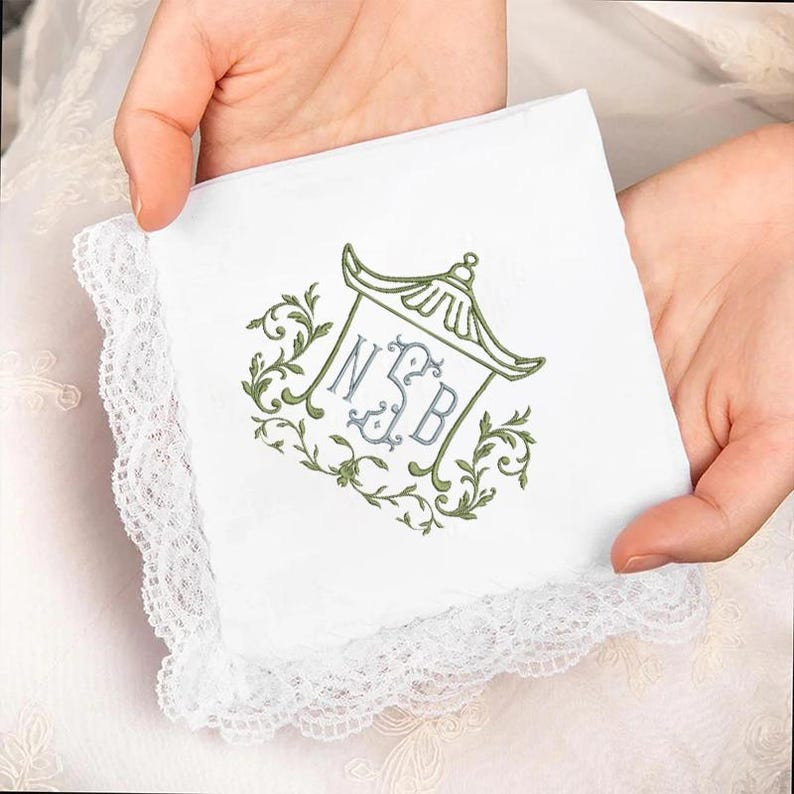 May include: A white square handkerchief featuring a delicate lace border and an embroidered design in the center. The embroidery depicts a pagoda-style roof outline in green, surrounding the monogram letters NSB in light blue, with green vines.