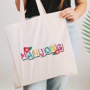 Puede incluir: Bolso tote de color crema con la palabra "MAHJONG" bordada en letras coloridas. Las letras est&aacute;n delineadas en negro y acentuadas con flores rosas y amarillas y hojas verdes. El bolso tiene dos asas.