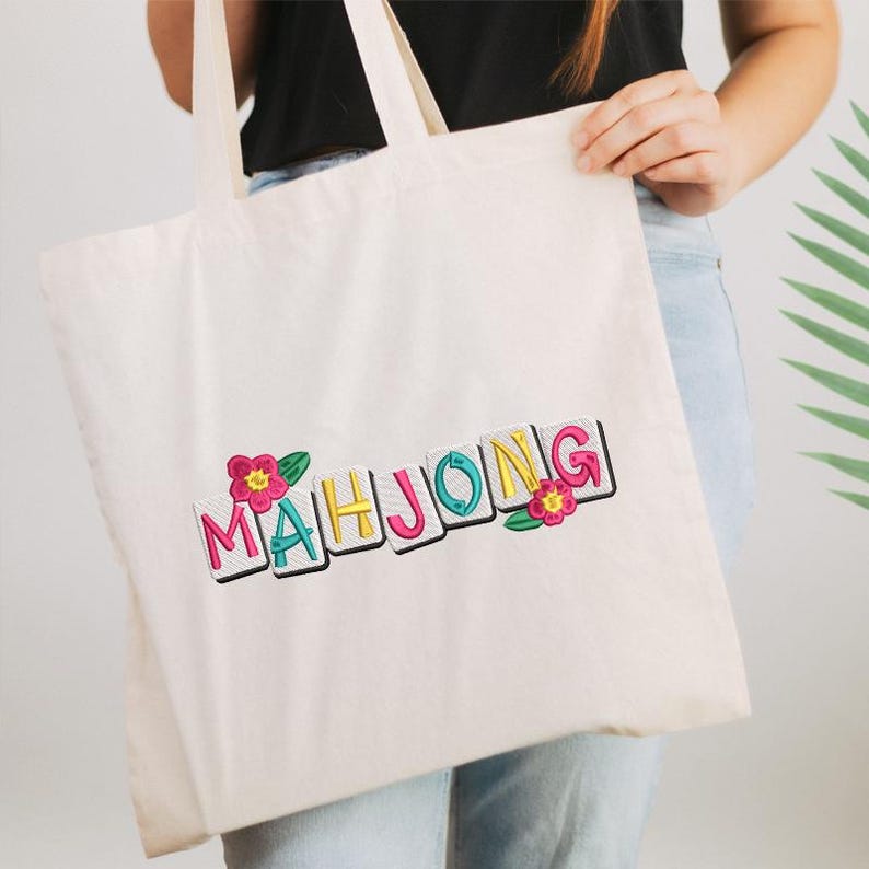 Puede incluir: Bolso tote de color crema con la palabra "MAHJONG" bordada en letras coloridas. Las letras est&aacute;n delineadas en negro y acentuadas con flores rosas y amarillas y hojas verdes. El bolso tiene dos asas.