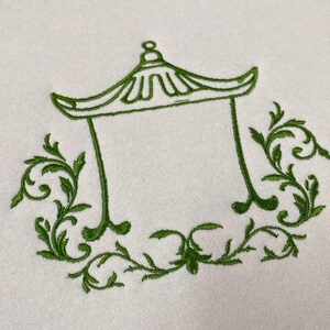 May include: A light-colored fabric featuring a green embroidered design of a pagoda-style roof structure. The central architectural outline is flanked and framed by intricate, symmetrical green leafy scrollwork and vine-like patterns.