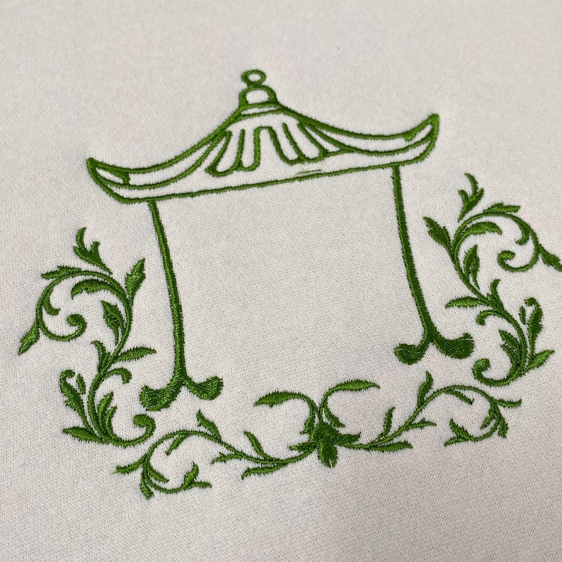 May include: A light-colored fabric featuring a green embroidered design of a pagoda-style roof structure. The central architectural outline is flanked and framed by intricate, symmetrical green leafy scrollwork and vine-like patterns.