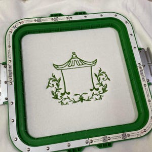 May include: A square green embroidery hoop with white magnetic strips holds a piece of white fabric. Centered on the fabric is a dark green machine-embroidered design featuring a pagoda-style roof structure surrounded by ornate scrollwork.