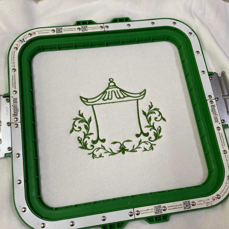 May include: A square green embroidery hoop with white magnetic strips holds a piece of white fabric. Centered on the fabric is a dark green machine-embroidered design featuring a pagoda-style roof structure surrounded by ornate scrollwork.