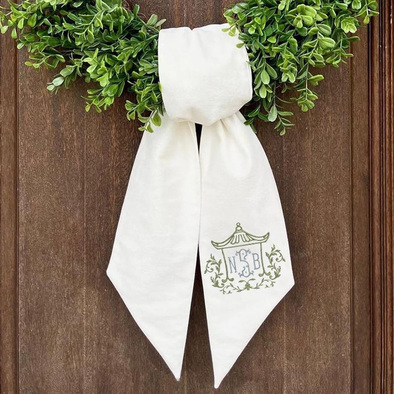 May include: A white fabric ribbon tied in a bow, featuring an embroidered green pagoda-style frame surrounding the monogram letters NSB. The ribbon is positioned over a circular green wreath with small leaves, creating a decorative accent piece.