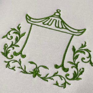 May include: An off-white fabric featuring an embroidered green design of a pagoda-style roof structure. The central architectural outline is flanked on both sides by intricate, symmetrical green leafy vine patterns that curve along the base.