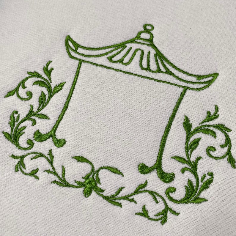 May include: An off-white fabric featuring an embroidered green design of a pagoda-style roof structure. The central architectural outline is flanked on both sides by intricate, symmetrical green leafy vine patterns that curve along the base.